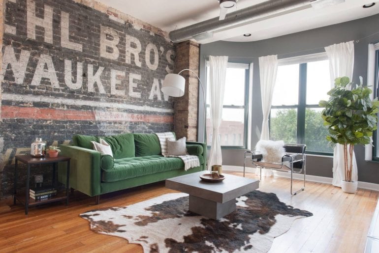 The living space has a painted brick wall, faux cowhide rug, large windows, and a plush green couch. 