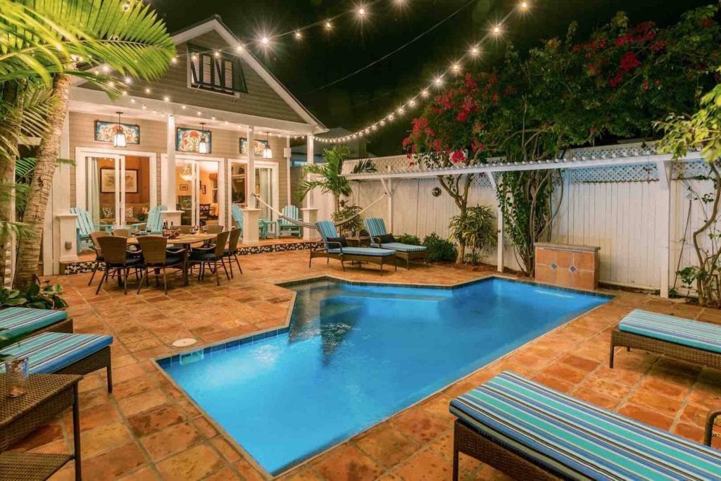 Backyard pavilion with in-ground pool, lounge chairs, and dining table lit by string lights.