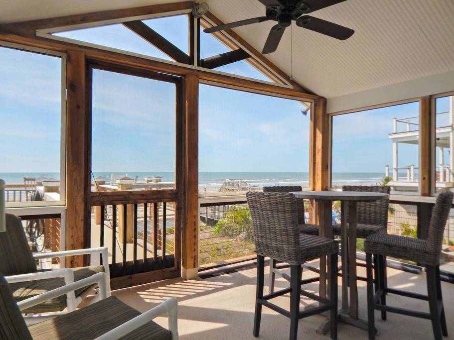 Screened-in porch features tall ceilings, a fan, a table with chairs, and a gorgeous beach view